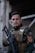 A person dressed in camouflage military attire is holding a black firearm. They are wearing a tactical vest with multiple pockets and a patch on the shoulder. The setting appears to be outdoors, possibly near a structure with metallic elements.