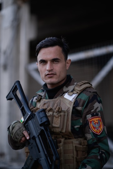 A person dressed in camouflage military attire is holding a black firearm. They are wearing a tactical vest with multiple pockets and a patch on the shoulder. The setting appears to be outdoors, possibly near a structure with metallic elements.