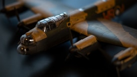 A detailed model of a vintage military aircraft with a canopy and distinctive propellers is captured in soft focus. The model features camouflage paint and intricate details on the wings and body.