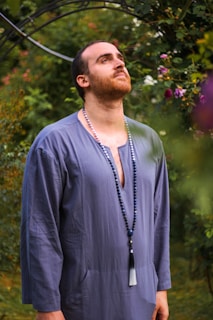 A person with a contemplative expression wearing a long, blue tunic and a beaded necklace stands in a lush garden setting, with greenery and purple flowers surrounding. The background includes a metal archway among the foliage.