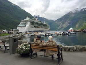 Two elderly people are sitting on a bench next to a body of water in a scenic area. They are wearing jackets and appear to be enjoying the view of a large cruise ship docked nearby. There are smaller boats and a pier in the background, with lush green mountains and misty clouds surrounding the area. A trash can and flower pot can be seen next to the bench.