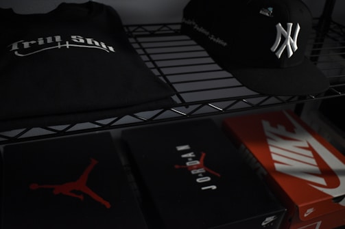 On a metal shelf, a black T-shirt with 'Trill Shit' printed in white sits beside a black baseball cap featuring a white emblem. Below, there are three shoeboxes; two are branded with 'Jordan' and 'Nike' logos.