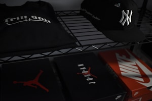 On a metal shelf, a black T-shirt with 'Trill Shit' printed in white sits beside a black baseball cap featuring a white emblem. Below, there are three shoeboxes; two are branded with 'Jordan' and 'Nike' logos.