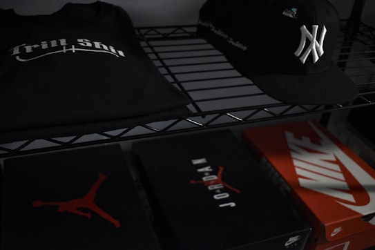 On a metal shelf, a black T-shirt with 'Trill Shit' printed in white sits beside a black baseball cap featuring a white emblem. Below, there are three shoeboxes; two are branded with 'Jordan' and 'Nike' logos.