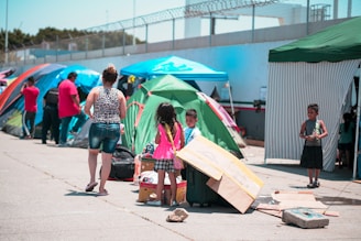A group of people are gathered in an outdoor area with several colorful tents. Children and adults are interacting, with some children playing near boxes and luggage. The scene includes temporary shelters and other camping equipment.