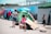 A group of people are gathered in an outdoor area with several colorful tents. Children and adults are interacting, with some children playing near boxes and luggage. The scene includes temporary shelters and other camping equipment.