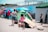 A group of people are gathered in an outdoor area with several colorful tents. Children and adults are interacting, with some children playing near boxes and luggage. The scene includes temporary shelters and other camping equipment.