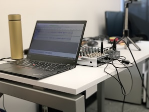 A laptop is placed on a white desk next to a golden thermos. The screen displays audio editing software. On the desk is also an audio mixer connected with various cables, suggesting a workspace for audio or music production. In the background, there are additional electronic devices and wires.