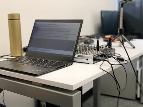 A laptop is placed on a white desk next to a golden thermos. The screen displays audio editing software. On the desk is also an audio mixer connected with various cables, suggesting a workspace for audio or music production. In the background, there are additional electronic devices and wires.