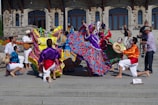 A vibrant group of performers in colorful traditional attire with face paint are dancing energetically on steps in front of a historical stone building. The performers are wearing long, flowing dresses adorned with bright floral patterns and lively shirts with sashes. Some musicians are playing instruments, adding to the festive atmosphere, and a cameraman captures the event.