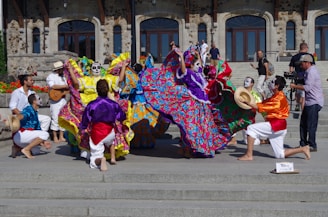 A vibrant group of performers in colorful traditional attire with face paint are dancing energetically on steps in front of a historical stone building. The performers are wearing long, flowing dresses adorned with bright floral patterns and lively shirts with sashes. Some musicians are playing instruments, adding to the festive atmosphere, and a cameraman captures the event.