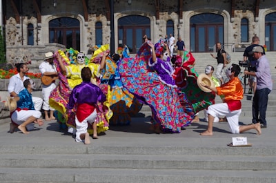 A vibrant group of performers in colorful traditional attire with face paint are dancing energetically on steps in front of a historical stone building. The performers are wearing long, flowing dresses adorned with bright floral patterns and lively shirts with sashes. Some musicians are playing instruments, adding to the festive atmosphere, and a cameraman captures the event.