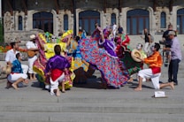 A vibrant group of performers in colorful traditional attire with face paint are dancing energetically on steps in front of a historical stone building. The performers are wearing long, flowing dresses adorned with bright floral patterns and lively shirts with sashes. Some musicians are playing instruments, adding to the festive atmosphere, and a cameraman captures the event.