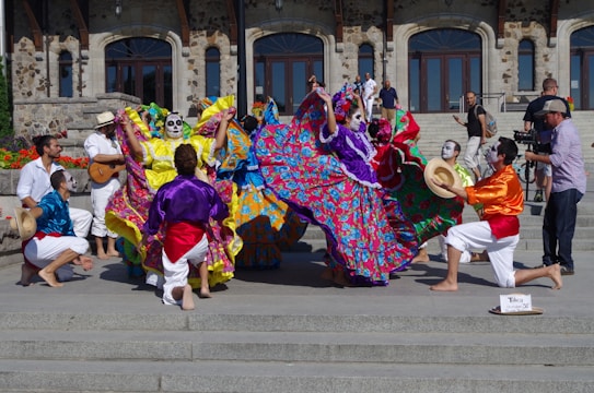 A vibrant group of performers in colorful traditional attire with face paint are dancing energetically on steps in front of a historical stone building. The performers are wearing long, flowing dresses adorned with bright floral patterns and lively shirts with sashes. Some musicians are playing instruments, adding to the festive atmosphere, and a cameraman captures the event.