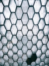 A pattern of hexagonal glass blocks forms a geometric texture. The translucent blocks are arranged tightly together, with some having visible imperfections and slight color variations. The surface reflects light, creating a mix of shadows and highlights.