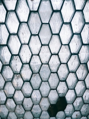 A pattern of hexagonal glass blocks forms a geometric texture. The translucent blocks are arranged tightly together, with some having visible imperfections and slight color variations. The surface reflects light, creating a mix of shadows and highlights.