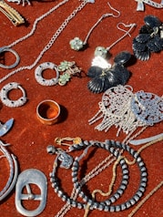 A collection of jewelry and accessories spread on a red velvet surface. Various items include earrings, rings, necklaces, and bracelets in different designs and colors such as silver, gold, and black. Some pieces have gemstone embellishments, while others have intricate metalwork.