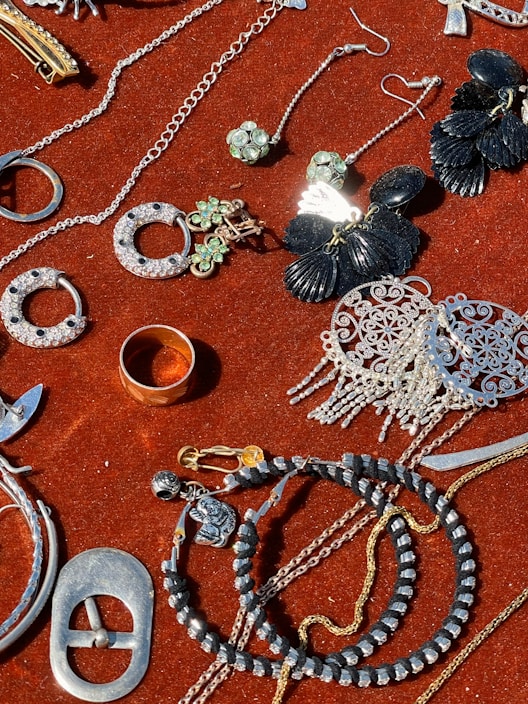 A collection of jewelry and accessories spread on a red velvet surface. Various items include earrings, rings, necklaces, and bracelets in different designs and colors such as silver, gold, and black. Some pieces have gemstone embellishments, while others have intricate metalwork.