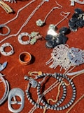 A collection of jewelry and accessories spread on a red velvet surface. Various items include earrings, rings, necklaces, and bracelets in different designs and colors such as silver, gold, and black. Some pieces have gemstone embellishments, while others have intricate metalwork.