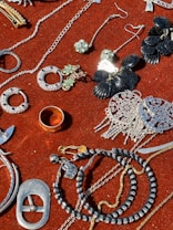 A collection of jewelry and accessories spread on a red velvet surface. Various items include earrings, rings, necklaces, and bracelets in different designs and colors such as silver, gold, and black. Some pieces have gemstone embellishments, while others have intricate metalwork.
