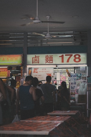 A busy market scene with people standing in line at a food stall, illuminated by overhead lighting. The stall displays a large sign featuring Chinese characters and the number 178. Various colorful posters and menus with prices are visible in the background.