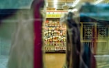 A display cabinet filled with numerous trophies is visible through a window, with reflections and blurred objects in the foreground. The trophies are arranged neatly and are surrounded by some decorative items. The setting appears to be indoors, with overhead lighting casting a warm glow.