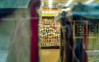 A display cabinet filled with numerous trophies is visible through a window, with reflections and blurred objects in the foreground. The trophies are arranged neatly and are surrounded by some decorative items. The setting appears to be indoors, with overhead lighting casting a warm glow.