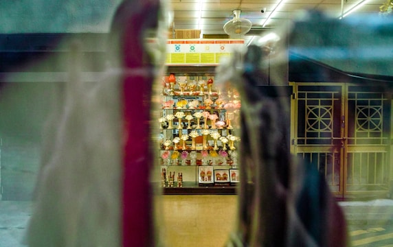 A display cabinet filled with numerous trophies is visible through a window, with reflections and blurred objects in the foreground. The trophies are arranged neatly and are surrounded by some decorative items. The setting appears to be indoors, with overhead lighting casting a warm glow.