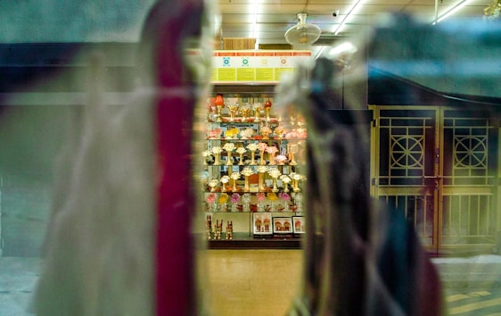 A display cabinet filled with numerous trophies is visible through a window, with reflections and blurred objects in the foreground. The trophies are arranged neatly and are surrounded by some decorative items. The setting appears to be indoors, with overhead lighting casting a warm glow.
