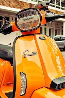A bright orange scooter featuring a hexagonal headlight with the brand name inscribed on it. The front panel displays 'Special V200' in stylized text. The scooter is parked in an outdoor setting with a partial view of a building in the background.