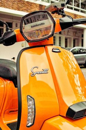 A bright orange scooter featuring a hexagonal headlight with the brand name inscribed on it. The front panel displays 'Special V200' in stylized text. The scooter is parked in an outdoor setting with a partial view of a building in the background.