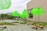 Several large, bright green structures resembling umbrellas or lampshades are placed on a paved outdoor area. They are mounted on wheeled bases surrounded by patches of grass. A modern building with a large rectangular opening stands in the background, and a few people can be seen walking nearby.