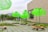 Several large, bright green structures resembling umbrellas or lampshades are placed on a paved outdoor area. They are mounted on wheeled bases surrounded by patches of grass. A modern building with a large rectangular opening stands in the background, and a few people can be seen walking nearby.