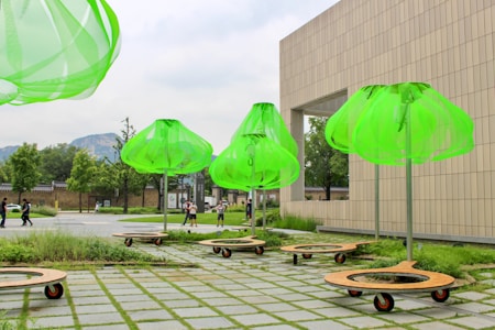 Several large, bright green structures resembling umbrellas or lampshades are placed on a paved outdoor area. They are mounted on wheeled bases surrounded by patches of grass. A modern building with a large rectangular opening stands in the background, and a few people can be seen walking nearby.