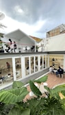 A modern, open-air café with two levels. The upper level has a white roof and features people seated at tables surrounded by potted plants. The lower level, enclosed with glass, has individuals working and socializing in a bright, minimalistic setting. Lush green foliage fills the foreground, adding a natural touch against the contemporary architecture. The atmosphere appears relaxed yet lively under a cloudy sky.