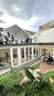 A modern, open-air café with two levels. The upper level has a white roof and features people seated at tables surrounded by potted plants. The lower level, enclosed with glass, has individuals working and socializing in a bright, minimalistic setting. Lush green foliage fills the foreground, adding a natural touch against the contemporary architecture. The atmosphere appears relaxed yet lively under a cloudy sky.