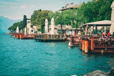 A picturesque lakeside scene with vibrant blue water meeting a row of waterside cafes adorned with flowering plants. Trees line the waterfront, leading to rustic buildings with charming architecture. Mountains are visible in the background, suggesting a serene and inviting atmosphere.