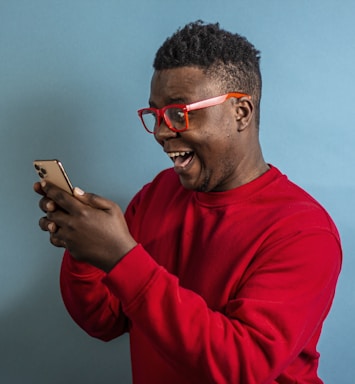 A person wearing bright red glasses and a matching red sweater is joyfully looking at a smartphone in their hands against a solid teal background. They have a wide, excited smile on their face.