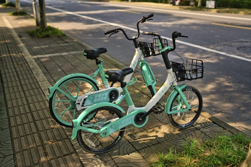 Two mint green and white rental bicycles are parked on a sidewalk next to a quiet road. Each bike is equipped with a front basket and has black seats and handlebars. The pavement is made up of patterned concrete tiles, and there is a patch of grass visible nearby. Sunlight casts soft shadows on the ground, suggesting a warm day.