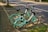 Two mint green and white rental bicycles are parked on a sidewalk next to a quiet road. Each bike is equipped with a front basket and has black seats and handlebars. The pavement is made up of patterned concrete tiles, and there is a patch of grass visible nearby. Sunlight casts soft shadows on the ground, suggesting a warm day.