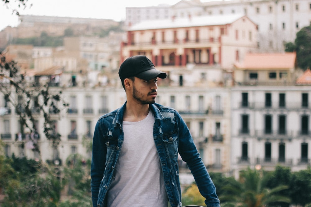 man in blue denim jacket and black cap standing near building during daytime