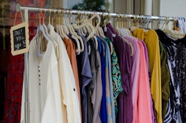 A clothing rack filled with various colorful garments, including dresses and shirts on hangers. A small blackboard sign indicates a sale with prices and discounts.
