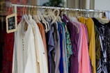A clothing rack filled with various colorful garments, including dresses and shirts on hangers. A small blackboard sign indicates a sale with prices and discounts.