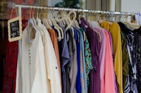 A clothing rack filled with various colorful garments, including dresses and shirts on hangers. A small blackboard sign indicates a sale with prices and discounts.