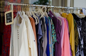 A clothing rack filled with various colorful garments, including dresses and shirts on hangers. A small blackboard sign indicates a sale with prices and discounts.