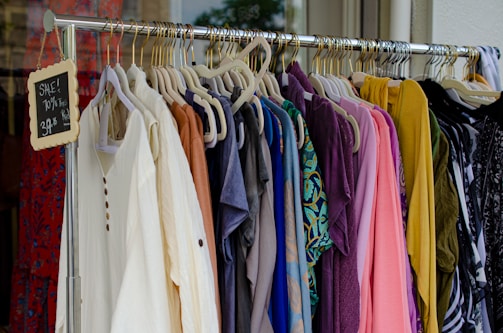 A clothing rack filled with various colorful garments, including dresses and shirts on hangers. A small blackboard sign indicates a sale with prices and discounts.