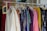 A clothing rack filled with various colorful garments, including dresses and shirts on hangers. A small blackboard sign indicates a sale with prices and discounts.