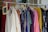 A clothing rack filled with various colorful garments, including dresses and shirts on hangers. A small blackboard sign indicates a sale with prices and discounts.