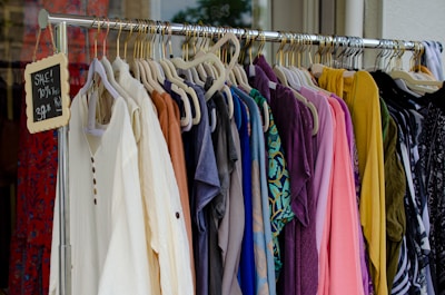 A clothing rack filled with various colorful garments, including dresses and shirts on hangers. A small blackboard sign indicates a sale with prices and discounts.