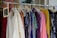 A clothing rack filled with various colorful garments, including dresses and shirts on hangers. A small blackboard sign indicates a sale with prices and discounts.